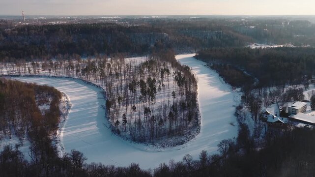 Aerial winter view shows frozen Gauja River curving around a snowy peninsula near Sigulda, Latvia, with Turaida tower on horizon, town, buildings, and long shadows.