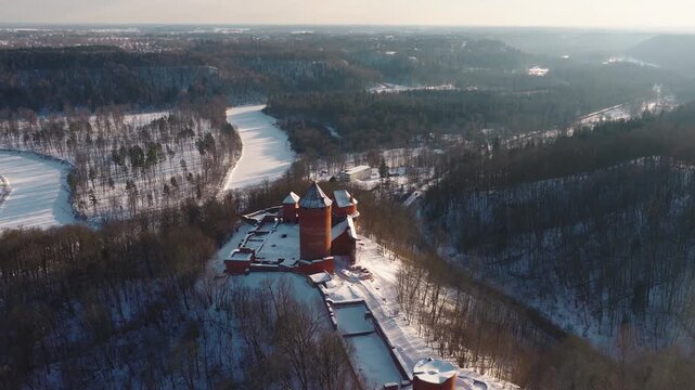 Aerial daylight sweep of Turaida Castle in Sigulda, Latvia, its red brick towers and walls dusted with snow as a drone tracks over the frozen Gauja River.