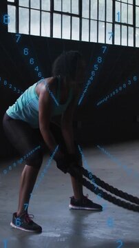 Woman doing battle-rope in warehouse gripping rope, driving waves as blue numbers tracking fitness