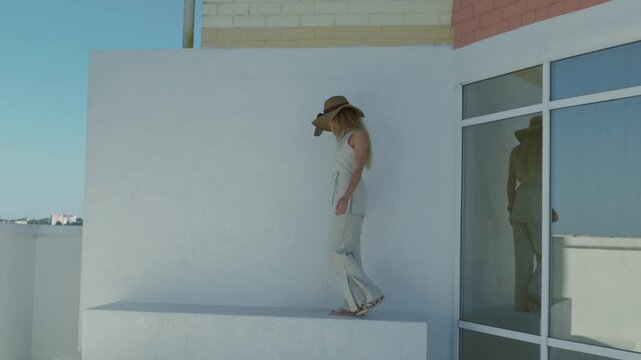 Woman in wide-brim hat walks along a ledge with modern architecture and city skyline
