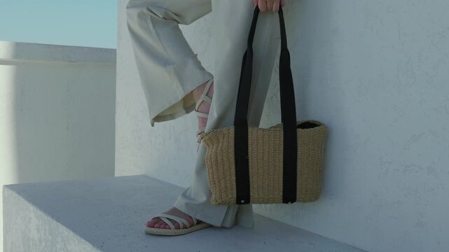 Woman in light linen pants and sandals holding a straw tote bag outdoors