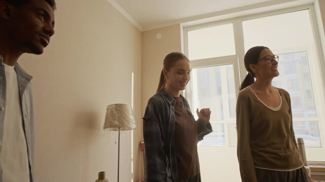 Group of four young multiethnic people standing among cardboard boxes and packed furniture in bright living room during housewarming celebration in new apartment, tracking medium shot