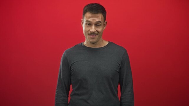 Hispanic man with mustache smirks with eyes closed in a red studio backdrop, facing forward; playful skepticism.