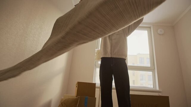 Low angle shot of young Caucasian man laying rug on floor while arranging interior during move into new apartment, cardboard boxes near window in background