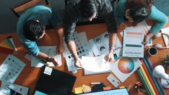 Top down aerial view of project manager looking at screen while designer writing idea. Smart business team explain idea of logo design while designer choose theme while looking at palette. Symposium.