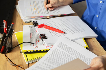 Professional editor reviews and marks up printed documents surrounded by stacks of papers, notebooks, and writing tools. Red marker corrections and annotations are visible on manuscript pages