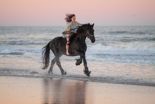 young woman riding Friesian horse stallion bare back on ocean beach seashore at dusk.
