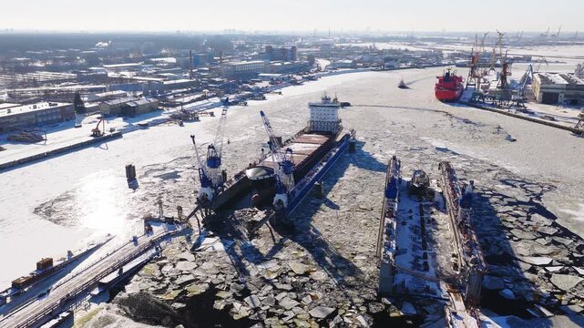 Riga Shipyard in Riga, Latvia shows a blue bulk carrier in a floating dock, cranes, long dry docks, a red tanker at the quay, broken ice, tugboats, and a cleared channel.