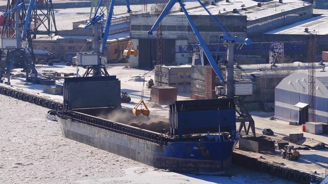 Aerial view of Riga Shipyard in Riga, Latvia shows a blue bulk carrier at the Daugava River quay as blue gantry cranes unload with a yellow grab amid ice and snow.