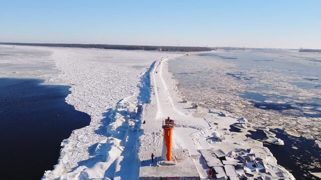 Aerial view shows bright orange Mangalsala lighthouse on a snow covered breakwater as pack ice drifts, deep water meets ice, and people walk in Riga, Latvia.