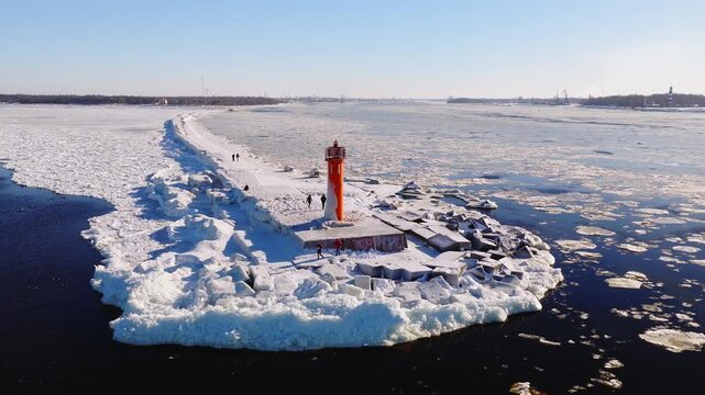 Aerial view shows bright orange Mangalsala lighthouse on a snow covered breakwater in Riga, Latvia. People walk on the icy jetty as ice floes drift on a clear winter day.