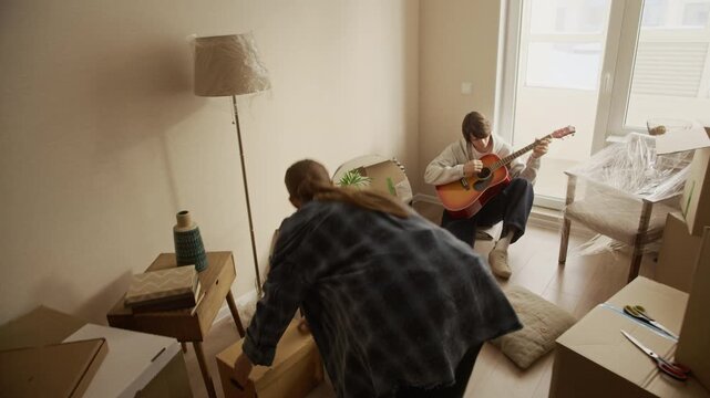 Tracking eye level shot of young Caucasian couple moving into new apartment, woman carrying cardboard boxes while man sitting on floor by window playing acoustic guitar in bright cozy living room