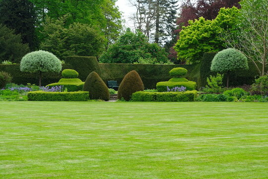 Manicured green lawn leads to a formal garden with ornate topiary and a secluded park bench against a dense hedge, embodying luxury landscaping, horticultural beauty, and serene estate elegance.