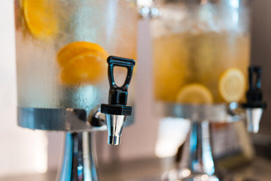 Refreshing beverages served from glass dispensers with taps for parties events. Large jars hold iced drinks with fruit slices like oranges, lemons. White cups stacked nearby for guests to use at