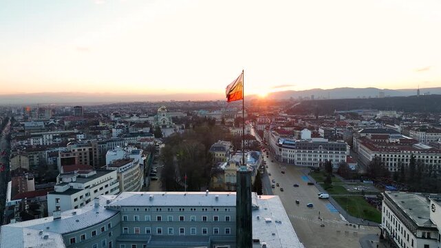 A close-up view of the flag of Bulgaria blowing in the wind against a sunset or sunrise over the capital city of Sofia. The Bulgarian flag flying over the Parliament building.