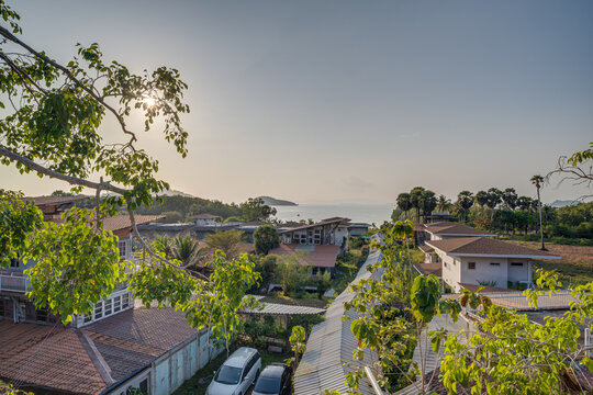 A ground-level view of a resort area featuring minimalist white buildings with thatched-style roofs. A narrow path leads through wild grass and tropical plants