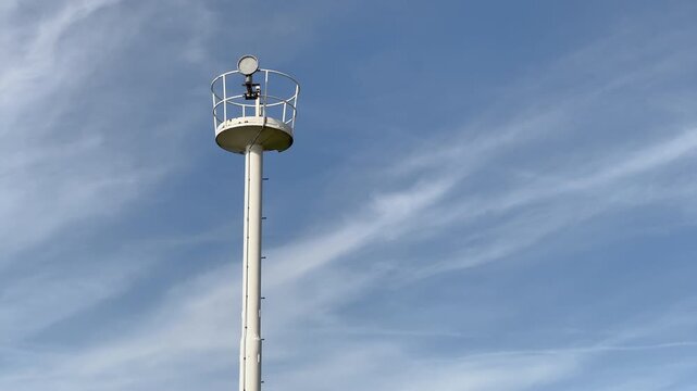 Close-up detail of weather station pole top featuring anemometer and navigation light mounted against blue sky with wispy clouds, meteorological equipment for coastal atmospheric monitoring.