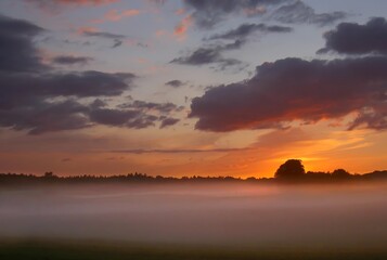 Obraz premium A dramatic sunset over a misty landscape, with the sun glowing orange on the horizon behind a layer of fog. This atmospheric scene captures the quiet transition from day to night in the countryside.