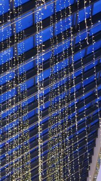 Blue atrium ceiling draped with lights, suspended vertical strings of warm white LEDs form shimmering curtain across modern mall, bold blue panels and dark beams create geometric rhythm, closeup