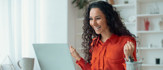 A woman with curly hair expresses joy while sitting at a desk. She is looking at her laptop and has...