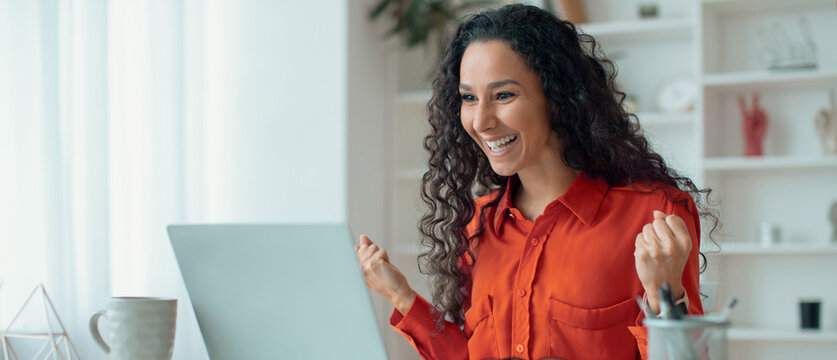 A woman with curly hair expresses joy while sitting at a desk. She is looking at her laptop and has her hands raised in celebration. The bright room has plants and organized items.