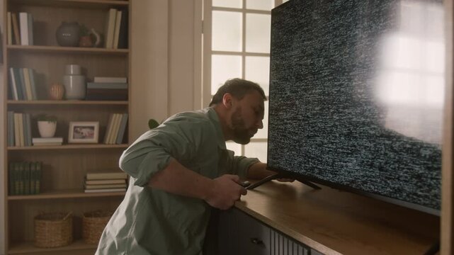Medium shot of young Caucasian man struggling to fix malfunctioning flat screen digital TV with static noise, examining remote control, checking cables and plugs to determine cause of breakdown