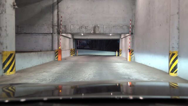 Point of view shot of a car driving out of a concrete underground parking garage at night, passing through an automatic opening barrier arm gate with flashing lights