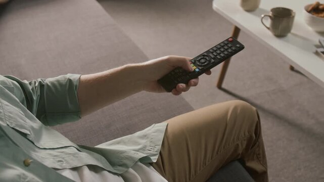 High-angle medium close-up of hand of unrecognizable male televiewer pressing button on remote control, while choosing movie, entertainment show or news channel on interactive menu on smart TV at home