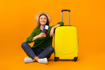 A woman sits on the floor with a yellow suitcase and travel documents. She wears a hat and smiles...