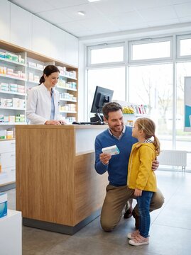 Father kneeling beside daughter in yellow jacket at pharmacy counter while pharmacist assists with prescription in bright, modern pharmacy interior