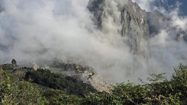 Machu Picchu, Peru - 26 June 2024. Clouds cover the ancient Inca site, obscuring terraces and stone buildings below a towering mountain, with dense greenery in front.