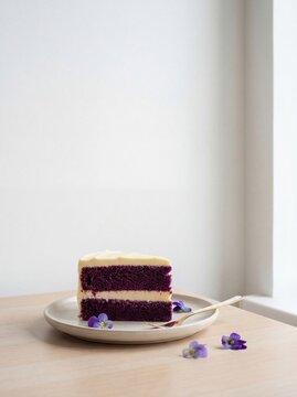 Layered purple cake slice with cream frosting on a beige plate, garnished with edible flowers, placed on a wooden table near a bright window