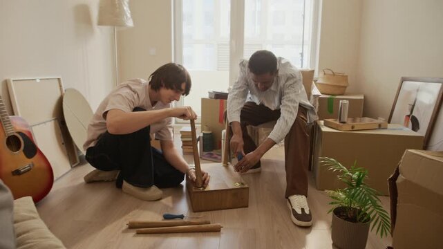 Dolly in shot of two women clinking champagne glasses while two men screwing wooden table legs during moving day in bright apartment among cardboard boxes and furniture