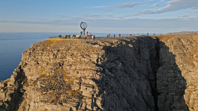 Drone aerial revealing the famous Nordkapp globe monument on the cliff edge in northern Norway. Dramatic Arctic cliffs rise above the Barents Sea.