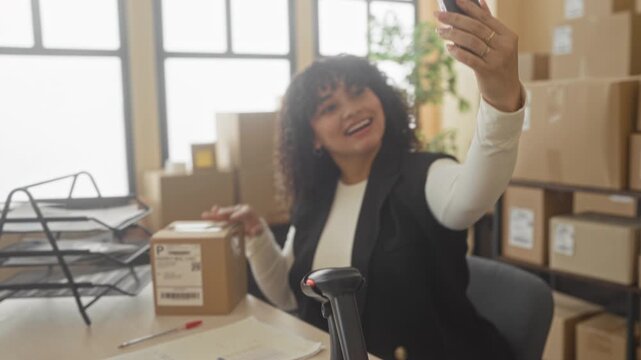 Woman holds smartphone and cardboard package, hand raised for selfie in a building shipping area; small business pride.