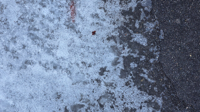 A top-down view of melting, slushy ice and frost encroaching upon a dark, damp asphalt surface. The texture is gritty and porous, showing cold transition and crystallization on the pavement.