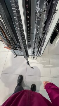 Buyer POV view. Overhead view of belt display with person in magenta jacket reaching for studded leather belt on vertical rack, chains and buckles visible on glossy