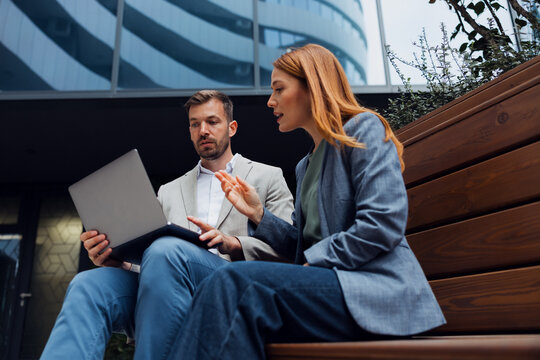 Professional colleagues discussing financial data on laptop outside a corporate headquarters