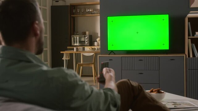 Medium rear shot of young man sitting on sofa in living room, switching channels with remote control while watching movie, show or sports match on TV with green screen with girlfriend or family