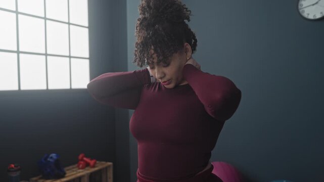 Woman stretching neck, hand on neck, tilting head in studio gym with dumbbells, exercise ball and window light; relief recovery.