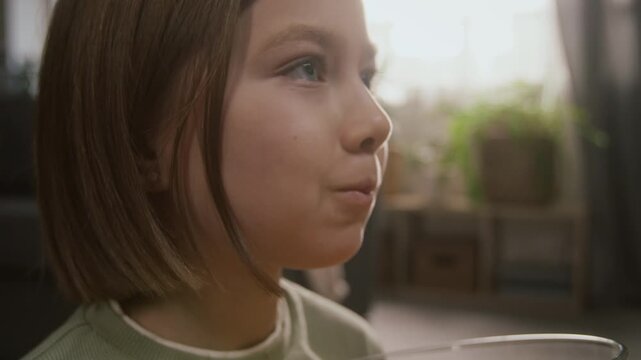 Close-up of face of young 8-year-old gen alpha girl munching potato chips from bowl, while watching cartoon or fun movie on TV at home, expressing attention and focus, kids entertainment concept
