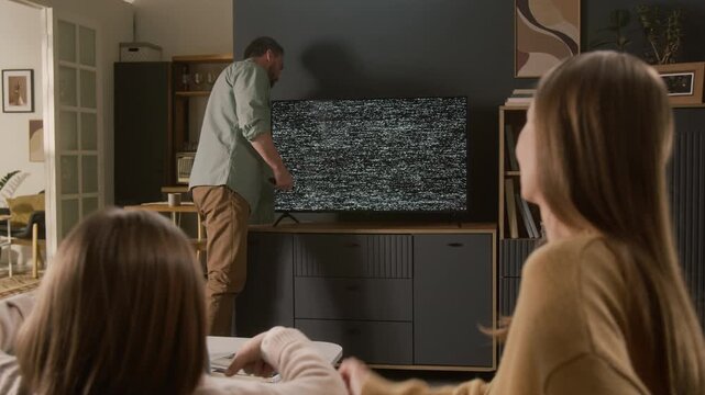 Medium full shot of annoyed Caucasian man standing by digital TV displaying gray static noise on screen, trying to set up with remote control, to watch movie with wife and daughter sitting on couch