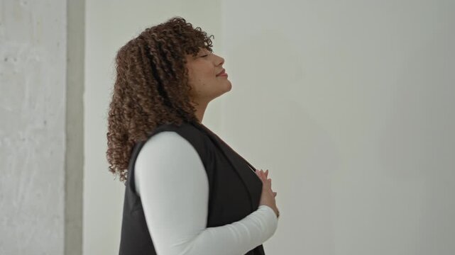 Woman with hands clasped to chest in an empty building, eyes closed and slight smile, wearing white long sleeve and black vest; serenity.