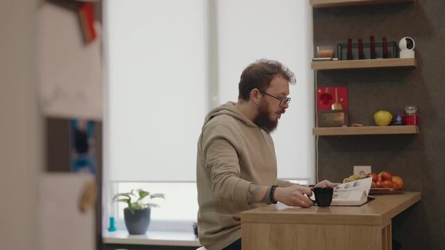 Bearded Man Enjoys Morning Coffee While Reading a Book in His Cozy Home Kitchen