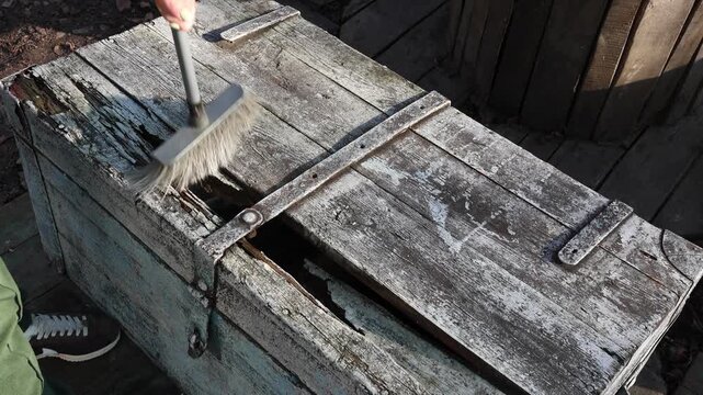 A person gently cleaning an old weathered wooden chest lid with a wire brush, getting it ready for a new coat of paint in the sunshine, the piece features a lock and rusty metal accents.