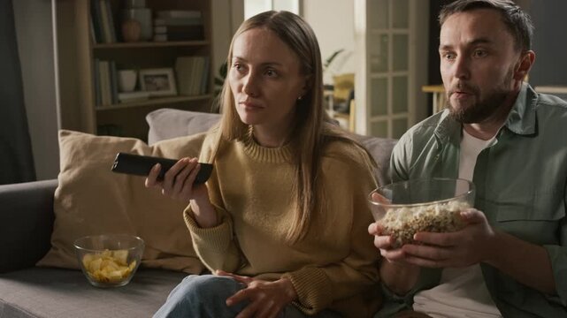 Medium zoom-in shot of scared young Caucasian couple watching slasher movie on TV, reacting with shock during violent suspense scene, jumping up with fear and spilling popcorn