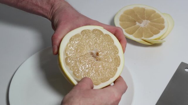 Hands masterfully peeling a halved pomelo to reveal its juicy segments, set against a clean white plate and a handy knife nearby for further use in the culinary creation.