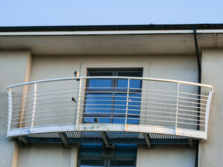 Fototapeta premium Curved balcony with white metal railings. Blue window reflects sky and clouds. Building facade shows slight wear and age. Ideal for urban living or architectural scenes.