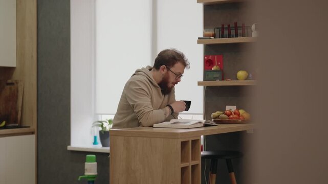 Bearded Man Enjoys Reading a Book and Drinking Coffee at Home in a Modern Kitchen
