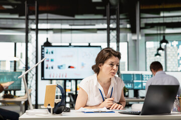 Fototapeta premium Caucasian female accountant works in office, focusing on improving funds for multinational company. Woman with pen in hand concentrates on project planning and managing business analytics on laptop.